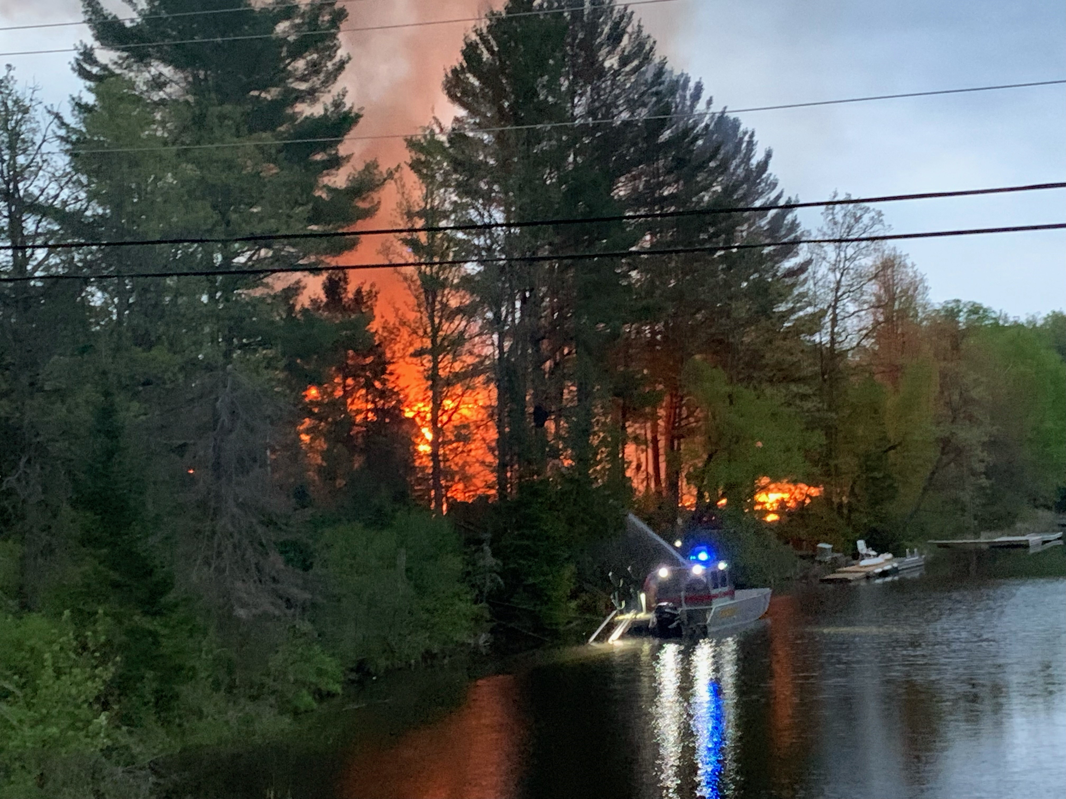 Flames from fire burning along the Shadow River with a fireboat in the water